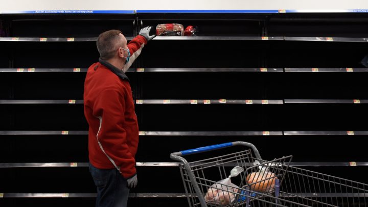 A man is wearing a medical mask at a grocery store and reaching for bread with empty shelves around him