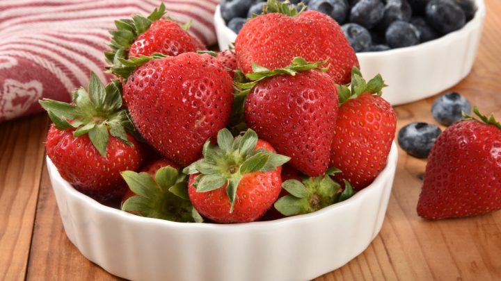 A bowl of fresh ripe strawberries with blueberries in the background.